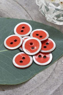 Boutons ronds à deux trous, finition émaillée orange corail vif cerclée d'un liseré blanc nacré, présentés en tas sur une feuille verte, avec une petite coupelle en verre facetté et des pétales de fleurs blanches en arrière-plan.