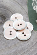 Two-hole round buttons, powder pink enamel finish rimmed with a pearly white trim, piled on a green leaf, with a small faceted glass dish and white flower petals in the background.