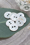 Boutons ronds à deux trous, finition émaillée blanc perlé cerclée d'un liseré blanc nacré, présentés en tas sur une feuille verte, avec une petite coupelle en verre facetté et des pétales de fleurs blanches en arrière-plan.