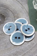 Boutons ronds à deux trous, finition émaillée bleu gris storm cerclée d'un liseré blanc nacré, présentés en tas sur une pierre grise, avec une feuille verte et une petite coupelle en verre facetté en arrière-plan.