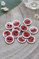 Boutons ronds à deux trous, finition émaillée rouge terracotta cerclée d'un liseré blanc nacré, présentés en tas sur une pierre grise, avec une feuille verte et une petite coupelle en verre facetté en arrière-plan.
