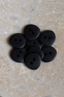 Seven small matte two-hole round buttons in matte black, piled in an oval marbled beige stone dish.