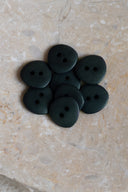 Seven small matte two-hole round buttons in very dark forest green, piled in an oval marbled beige stone dish.