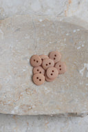 Sept petits boutons ronds mats à deux trous, coloris beige rosé sable, présentés en tas dans une coupelle en pierre ovale beige marbrée.