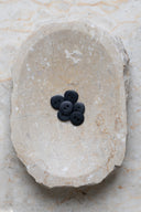 Sept petits boutons ronds mats à deux trous, coloris noir intense (night), présentés en tas dans une coupelle en pierre ovale beige marbrée.