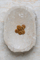 Sept petits boutons ronds mats à deux trous, coloris ocre miel, présentés en tas dans une coupelle en pierre ovale beige marbrée.