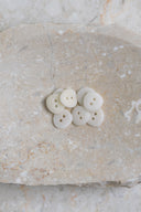 Sept petits boutons ronds mats à deux trous, coloris blanc cassé, présentés en tas dans une coupelle en pierre ovale beige marbrée.