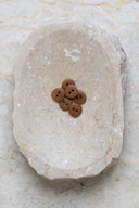 Sept petits boutons ronds mats à deux trous, coloris brun caramel pecan pie, présentés en tas dans une coupelle en pierre ovale beige marbrée.