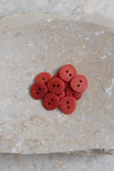 Sept petits boutons ronds mats à deux trous, coloris rouge orangé tangerine, présentés en tas dans une coupelle en pierre ovale beige marbrée.
