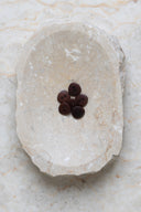 Sept petits boutons ronds mats à deux trous, coloris brun rouille foncé, présentés en tas dans une coupelle en pierre ovale beige marbrée.