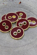 Five two-hole round buttons, glossy burgundy amaranth enamel finish, rimmed with a metallic gold trim, set on grey stone with scattered dried flower petals in the background.