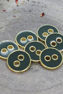 Five two-hole round buttons, glossy dark cedar green enamel finish, rimmed with a metallic gold trim, set on grey stone with scattered dried flower petals in the background.