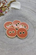 Five two-hole round buttons, glossy warm terracotta orange enamel finish, rimmed with a metallic gold trim, set on grey stone with scattered dried flower petals in the background.