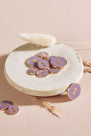 Boutons ronds à deux trous, finition émaillée brillante violet parme, cerclés d'un liseré métallique doré, présentés en tas sur une petite coupelle en céramique blanche, sur fond rose avec un épi de lagurus séché.