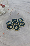 Sept boutons ronds à deux trous, finition émaillée brillante vert forêt très foncé, cerclés d'un liseré métallique doré, posés sur une pierre grise, avec un petit vase transparent et des pétales séchés en arrière-plan.