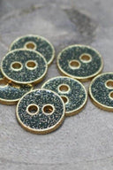 Seven small two-hole round buttons, gold glitter finish over a dark cedar green base, rimmed with a metallic gold trim, photographed close-up on grey stone.