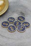 Sept petits boutons ronds à deux trous, finition pailletée dorée sur fond bleu cobalt, cerclés d'un liseré métallique doré, photographiés en gros plan sur une pierre grise avec une coupelle dorée floutée en arrière-plan.