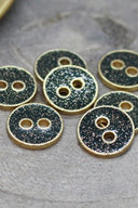 Seven small two-hole round buttons, gold glitter finish over a deep forest green base, rimmed with a metallic gold trim, photographed close-up on grey stone.