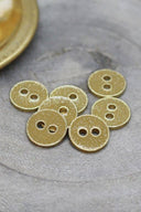 Seven small two-hole round buttons, gold glitter finish over a golden mustard yellow base, rimmed with a metallic gold trim, set on grey stone with a blurred gold dish in the background.