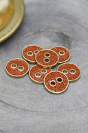 Sept petits boutons ronds à deux trous, finition pailletée dorée sur fond orange tangerine, cerclés d'un liseré métallique doré, posés sur une pierre grise, avec une coupelle dorée floutée en arrière-plan.