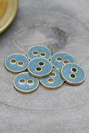 Sept petits boutons ronds à deux trous, finition pailletée dorée sur fond bleu-gris clair orageux, cerclés d'un liseré métallique doré, posés sur une pierre grise, avec une coupelle dorée floutée en arrière-plan.