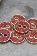 Sept petits boutons ronds à deux trous, finition pailletée dorée sur fond rouge terracotta, cerclés d'un liseré métallique doré, photographiés en gros plan sur une pierre grise.