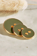 Trois boutons ronds à deux trous de tailles dégradées, finition émaillée brillante vert lierre foncé, cerclés d'un liseré métallique doré, posés sur une pierre claire avec un brin d'herbe séchée (lagurus) en arrière-plan.