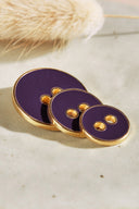 Trois boutons ronds à deux trous de tailles dégradées, finition émaillée brillante violet profond, cerclés d'un liseré métallique doré, posés sur une pierre claire avec un brin d'herbe séchée (lagurus) en arrière-plan.