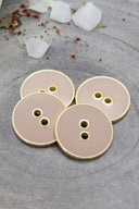 Cinq boutons ronds à deux trous, finition émaillée brillante beige rosé sable, cerclés d'un liseré métallique doré, posés sur une pierre grise avec des pétales de fleurs séchées éparpillés en arrière-plan.