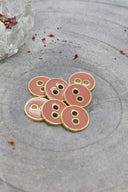 Seven two-hole round buttons, glossy peachy melba pink enamel finish, rimmed with a metallic gold trim, set on grey stone with a small clear vase and dried petals in the background.