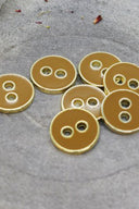 Cinq boutons ronds à deux trous, finition émaillée brillante ocre caramel, cerclés d'un liseré métallique doré, posés sur une pierre grise avec des pétales de fleurs séchées éparpillés en arrière-plan.