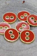 Cinq boutons ronds à deux trous, finition émaillée brillante rouge orangé vif, cerclés d'un liseré métallique doré, posés sur une pierre grise avec des pétales de fleurs séchées éparpillés en arrière-plan.