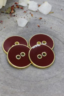 Cinq boutons ronds à deux trous, finition émaillée brillante brun rouille bordeaux, cerclés d'un liseré métallique doré, posés sur une pierre grise avec des pétales de fleurs séchées éparpillés en arrière-plan.