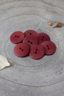Sept boutons ronds classiques à deux trous, effet bois veiné bordeaux amarante, présentés en tas sur une pierre grise, avec une feuille séchée en arrière-plan.