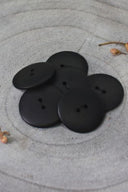 Sept boutons ronds classiques à deux trous, effet bois veiné noir mat profond, présentés en tas sur une pierre grise, avec une feuille séchée en arrière-plan.