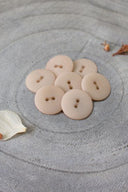 Sept boutons ronds classiques à deux trous, effet bois veiné rose pâle blush, présentés en tas sur une pierre grise, avec une feuille séchée en arrière-plan.