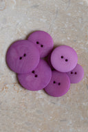 Cinq à sept boutons ronds classiques à deux trous, de tailles graduées, en résine effet bois veiné rose fuchsia vif bubble-gum, présentés en tas sur une dalle de pierre beige marbrée.