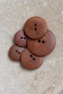 Cinq à sept boutons ronds classiques à deux trous, de tailles graduées, en résine effet bois veiné brun châtaigne rosé, présentés en tas sur une dalle de pierre beige marbrée.