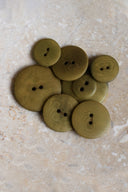 Cinq à sept boutons ronds classiques à deux trous, de tailles graduées, en résine effet bois veiné vert olive kaki (dreamy twilight), présentés en tas sur une dalle de pierre beige marbrée.