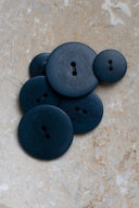 Cinq à sept boutons ronds classiques à deux trous, de tailles graduées, en résine effet bois veiné bleu-vert forêt très foncé, présentés en tas sur une dalle de pierre beige marbrée.
