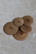 Cinq à sept boutons ronds classiques à deux trous, de tailles graduées, en résine effet bois veiné beige caramel glacé (iced mocha), présentés en tas sur une dalle de pierre beige marbrée.
