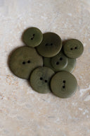 Cinq à sept boutons ronds classiques à deux trous, de tailles graduées, en résine effet bois veiné vert olive kaki, présentés en tas sur une dalle de pierre beige marbrée.