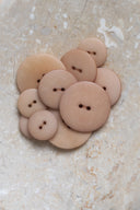Cinq à sept boutons ronds classiques à deux trous, de tailles graduées, en résine effet bois veiné beige rosé sable, présentés en tas sur une dalle de pierre beige marbrée.