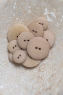 Cinq à sept boutons ronds classiques à deux trous, de tailles graduées, en résine effet bois veiné beige rosé argile clair (milky clay), présentés en tas sur une dalle de pierre beige marbrée.
