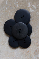 Cinq à sept boutons ronds classiques à deux trous, de tailles graduées, en résine effet bois veiné noir mat profond (night), présentés en tas sur une dalle de pierre beige marbrée.