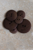 Five to seven classic two-hole round buttons, graduated sizes, in wood-grain-effect resin, chocolate brown, piled on a slab of marbled beige stone.