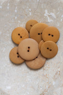 Cinq à sept boutons ronds classiques à deux trous, de tailles graduées, en résine effet bois veiné ocre miel, présentés en tas sur une dalle de pierre beige marbrée.