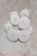 Cinq à sept boutons ronds classiques à deux trous, de tailles graduées, en résine effet bois veiné blanc cassé nacré, présentés en tas sur une dalle de pierre beige marbrée.