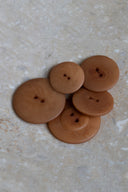 Cinq à sept boutons ronds classiques à deux trous, de tailles graduées, en résine effet bois veiné brun caramel pecan pie, présentés en tas sur une dalle de pierre beige marbrée.