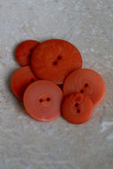 Cinq à sept boutons ronds classiques à deux trous, de tailles graduées, en résine effet bois veiné orange tangerine, présentés en tas sur une dalle de pierre beige marbrée.